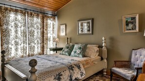 A bedroom with a wooden ceiling, a bed with floral bedding, two wall-mounted lamps, and a chair.