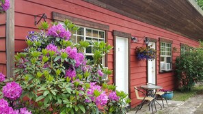 Une maison rouge avec une porte blanche, une fenêtre ornée d’un pot de fleurs suspendu et une petite table d’extérieur avec des chaises.
