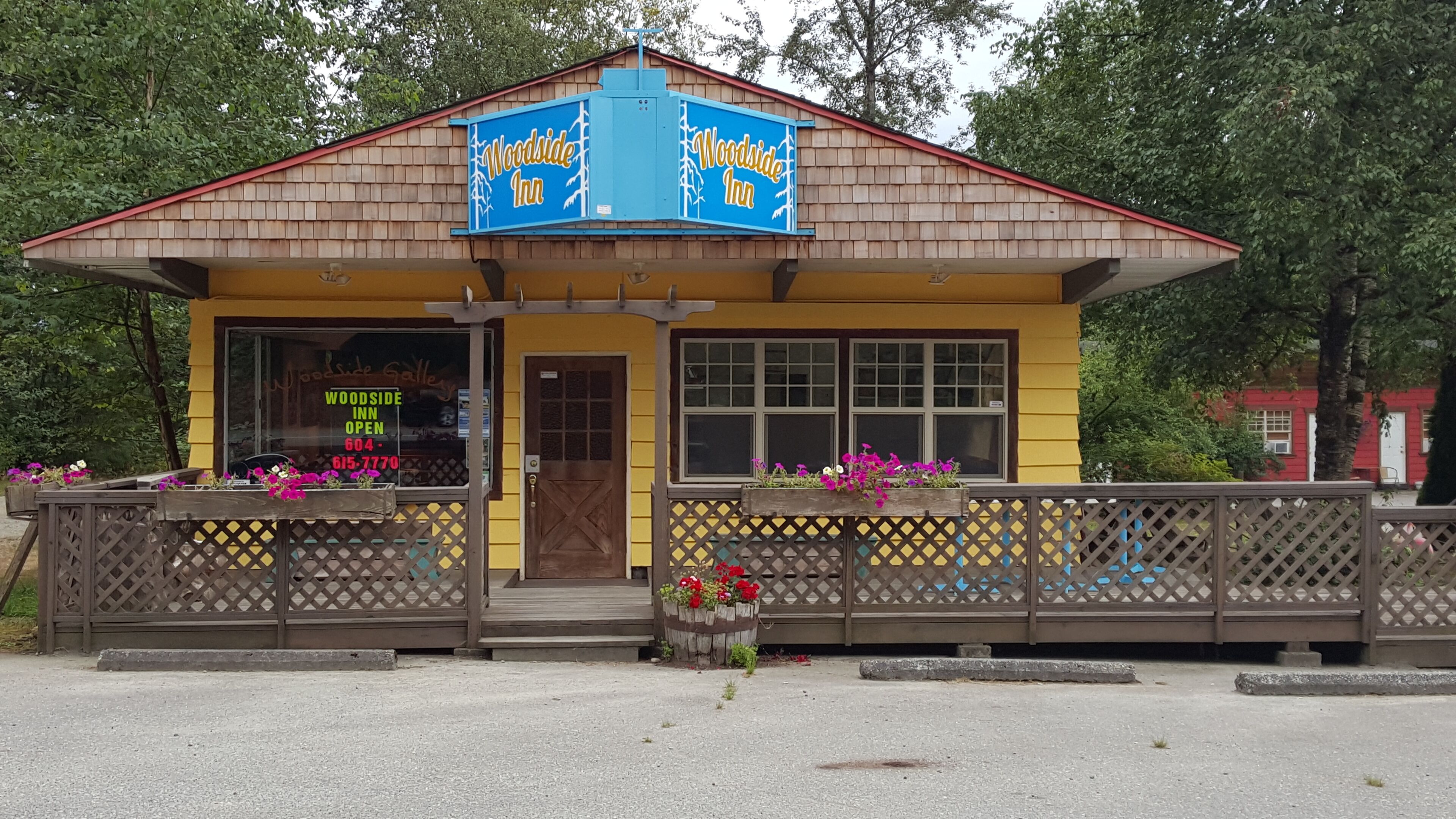 A small, single-story building with a yellow facade and a red roof, labeled "Woodside Inn." It features a wooden entrance with a glass door, a window with a sign, and a wooden fence with flower boxes.