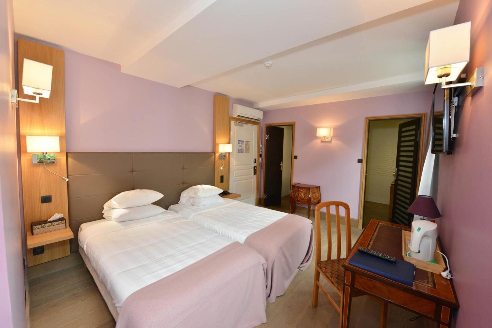 A hotel room with two beds, a desk with a chair, a table with a remote control, and a wall-mounted lamp.