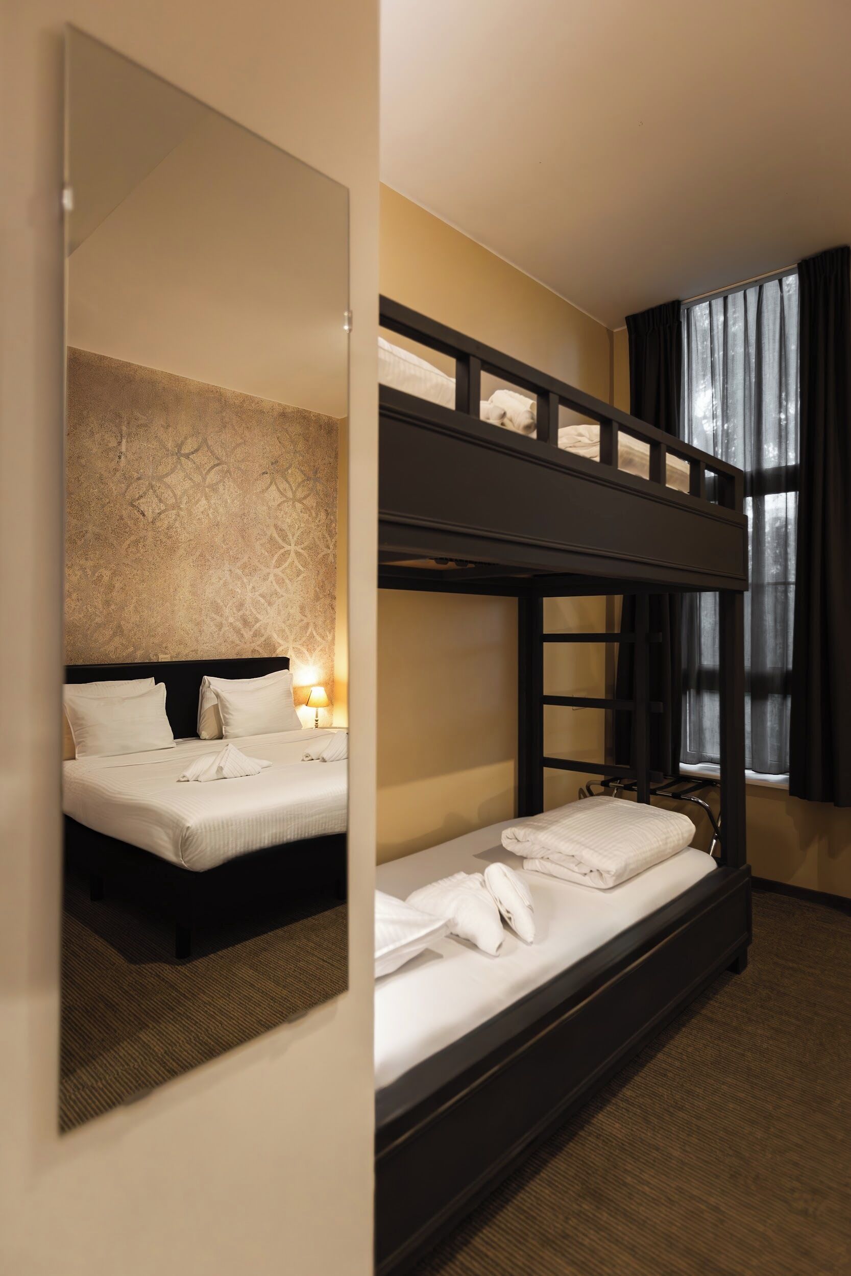 A hotel room with a bunk bed, a large mirror, and a window with curtains.