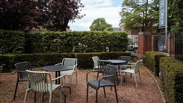 An outdoor seating area with tables and chairs, surrounded by hedges and trees.