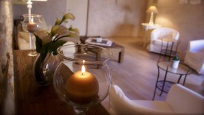 A cozy living room with a lit candle in a glass holder, a white sofa, a small round table with a plant, and a lamp on a side table. - Hôtel Le Crillon (Murs)