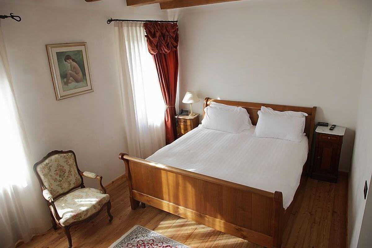 A bedroom with a wooden bed, a chair, a nightstand, a lamp, and a painting on the wall.