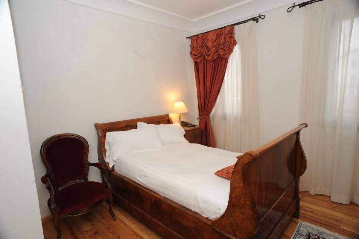 A single bed with a wooden frame and white bedding, a red upholstered chair, and a nightstand with a lamp.