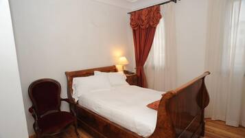 A single bed with a wooden frame and white bedding, a red upholstered chair, and a nightstand with a lamp.