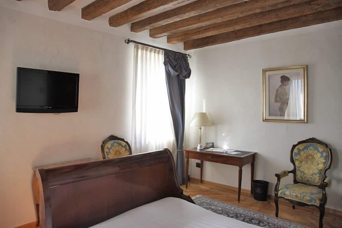 A bedroom with a wooden bed, a flat-screen TV mounted on the wall, a desk with a lamp, a chair, and a framed picture on the wall.