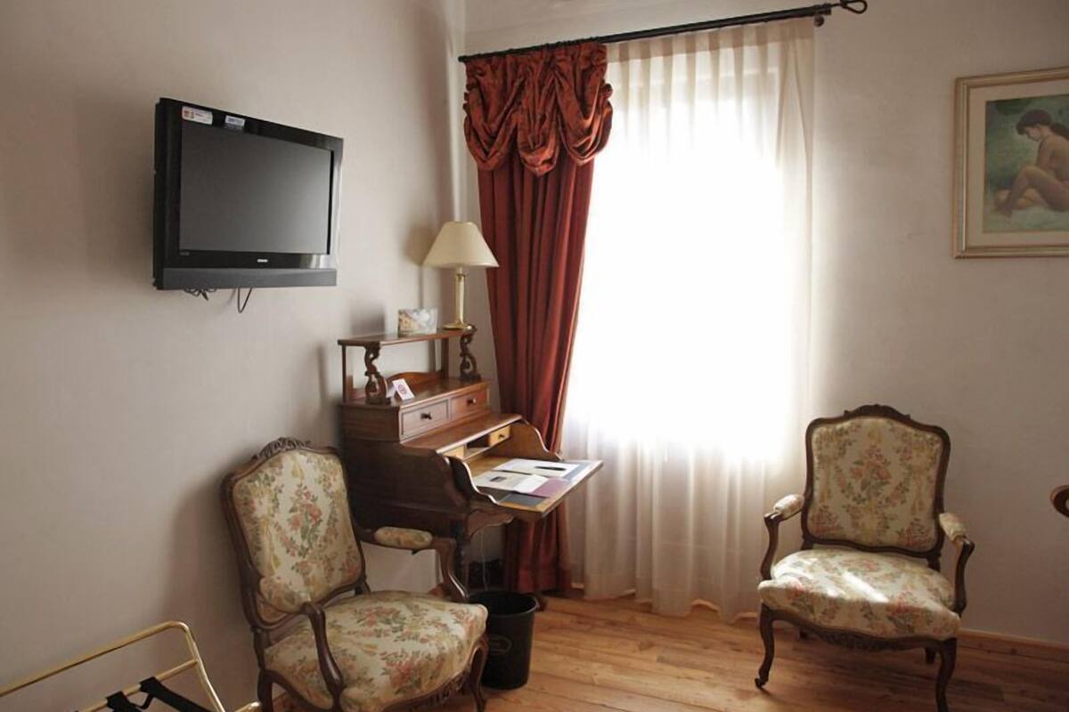 A room with a television mounted on the wall, a desk with a lamp, two floral armchairs, and a window with curtains.