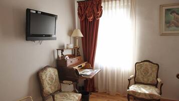 Une pièce avec un téléviseur fixé au mur, un bureau avec une lampe, deux fauteuils à motifs floraux et une fenêtre avec des rideaux.