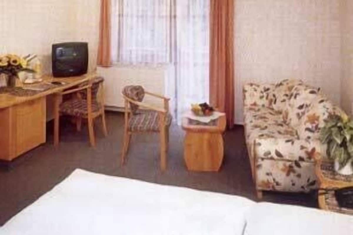 A small, furnished room with a wooden desk, a television, a patterned sofa, and a window with curtains.
