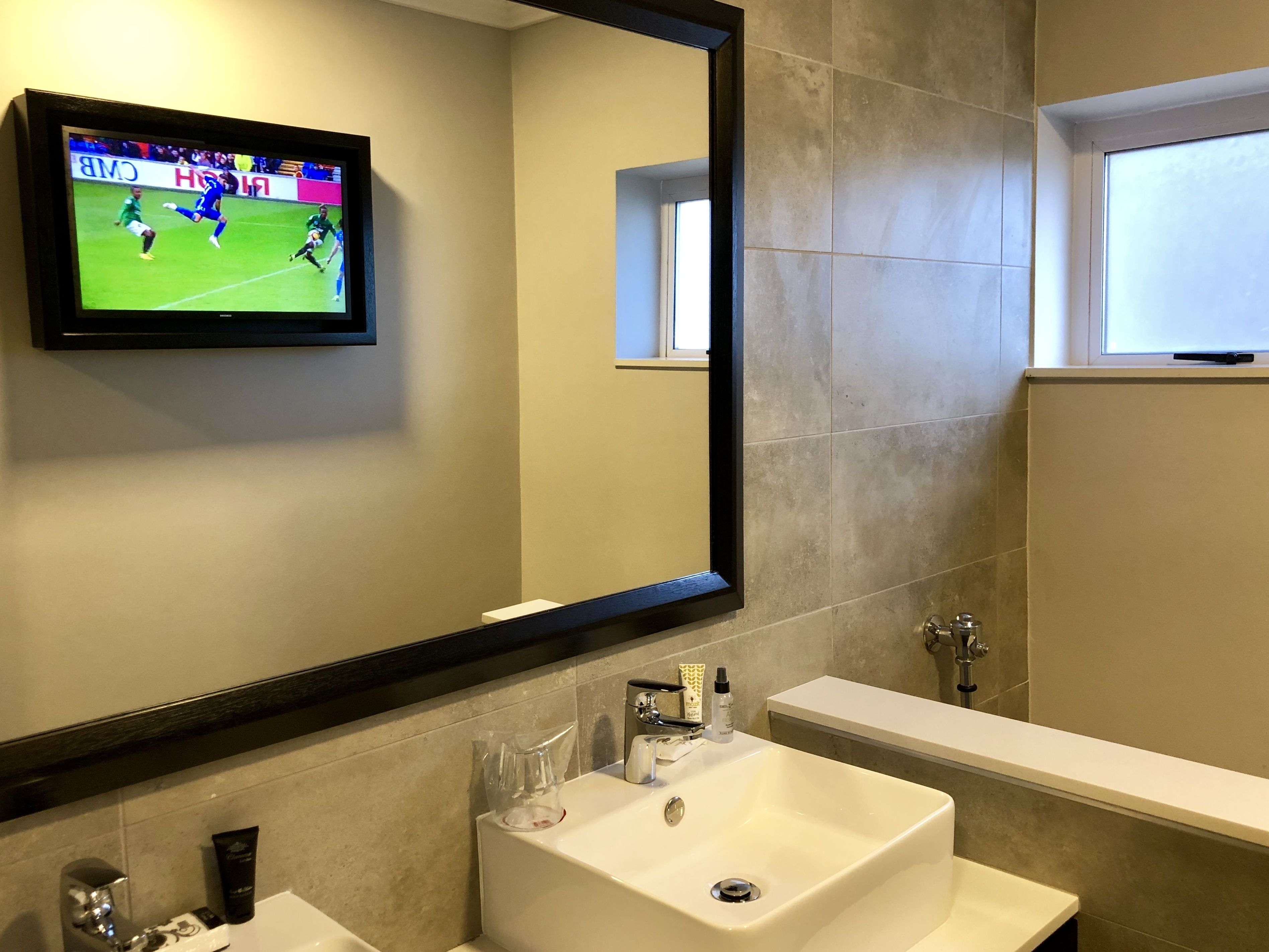 A bathroom with a large mirror, a sink, a toilet, and a television mounted on the wall displaying a sports game.