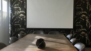 A conference room with a large wooden table, leather chairs, a projector screen, and a tropical leaf-patterned wall.
