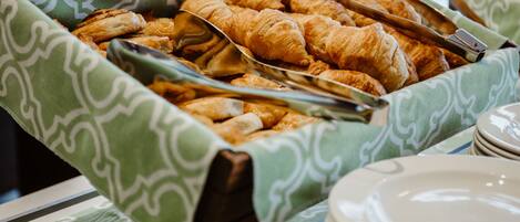 A buffet with a green patterned tablecloth containing croissants and other pastries, with tongs and plates nearby.
