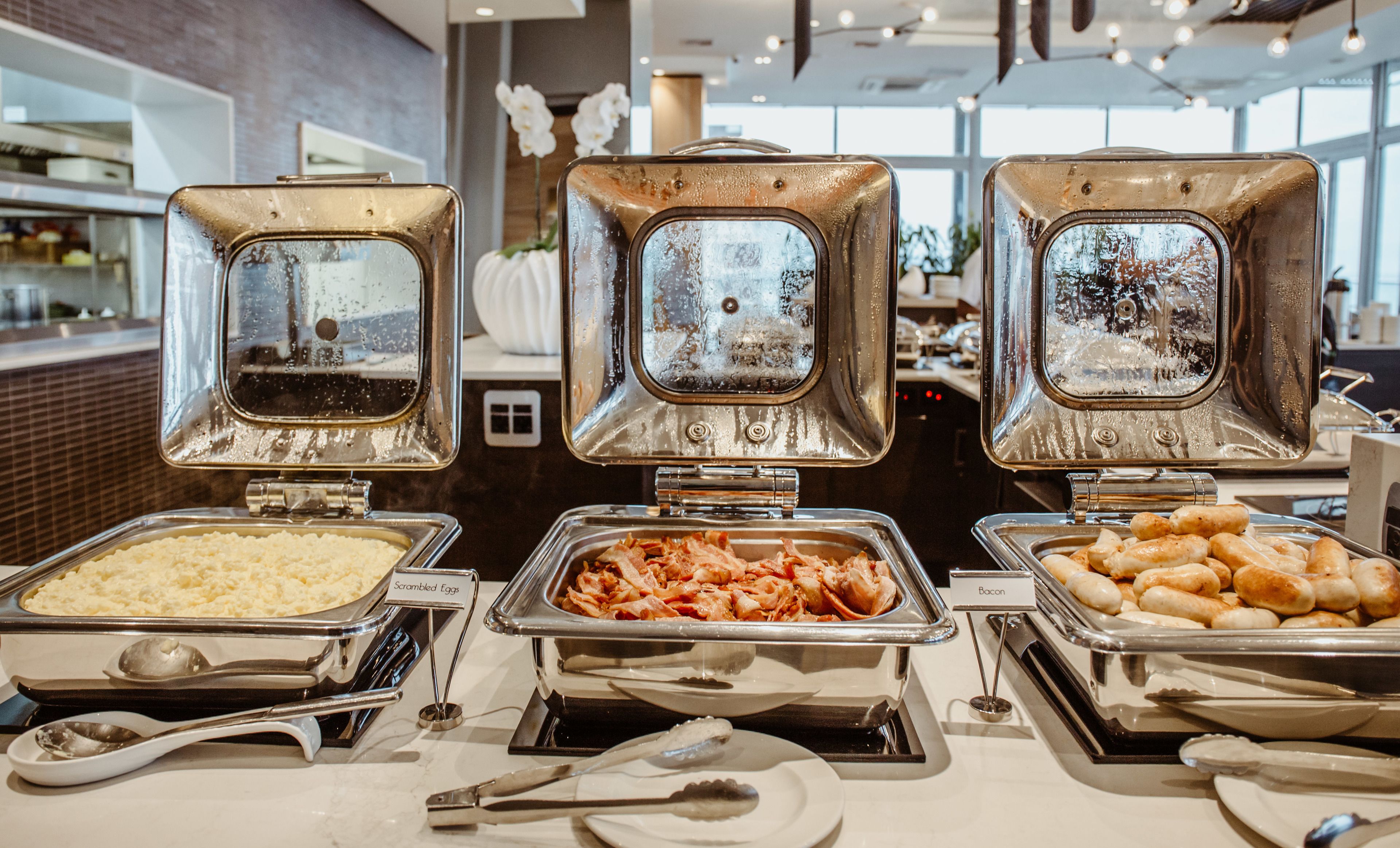 A buffet with labeled containers of scrambled eggs, bacon, and sausages.