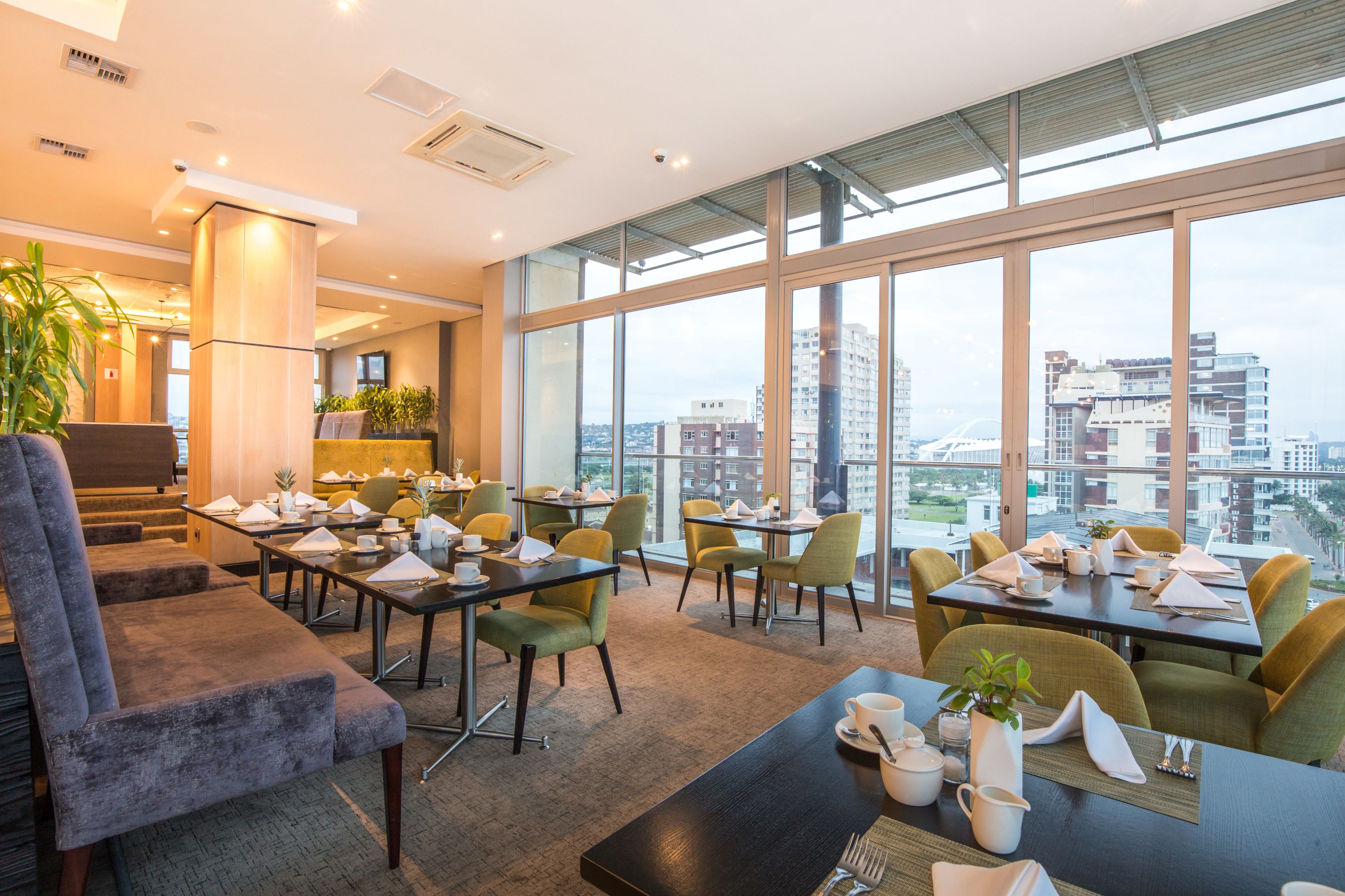 A modern hotel lobby with a high ceiling, large windows offering city views, and a mix of seating arrangements including tables and chairs.