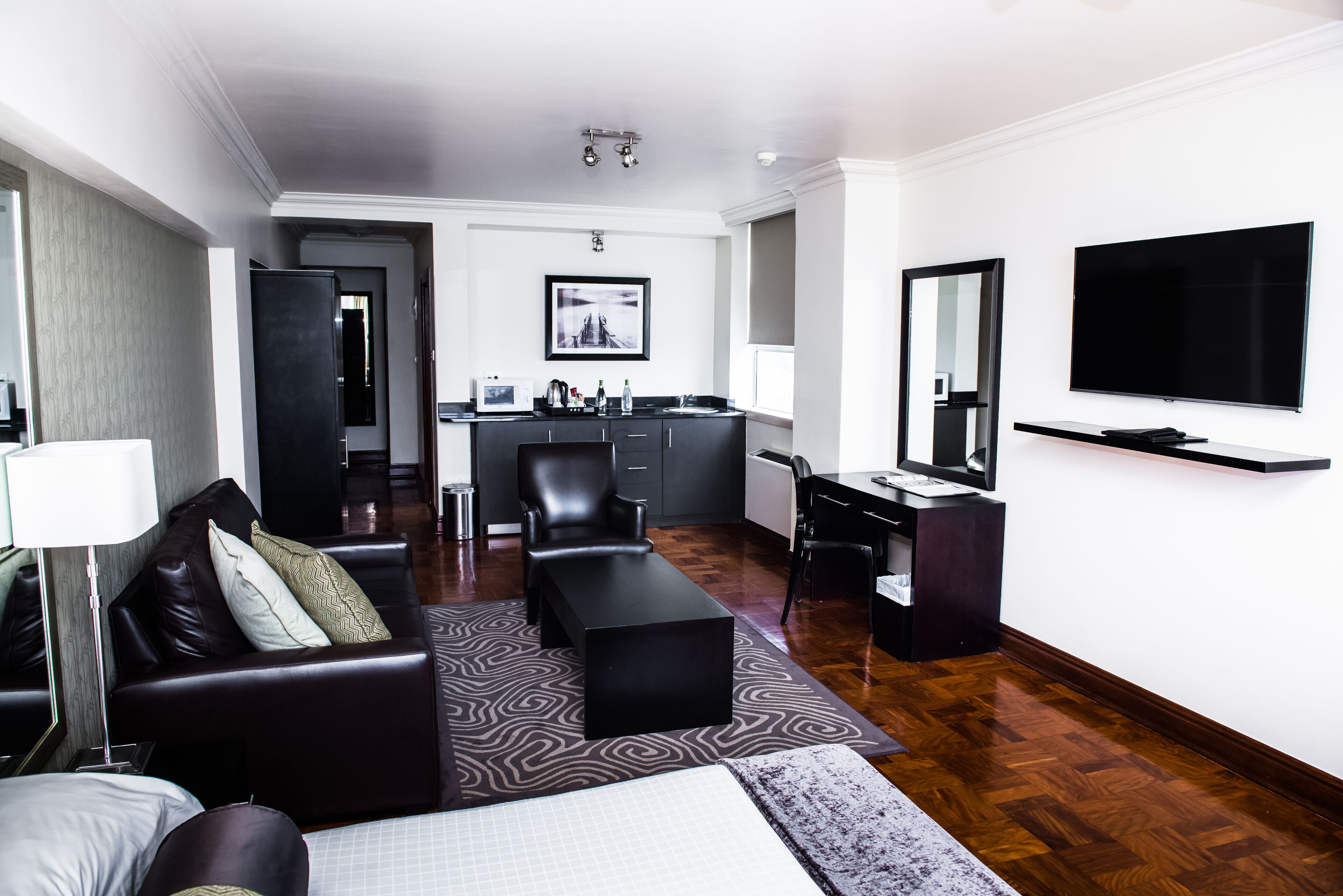 A modern hotel room with a bed, a sofa, a desk, a chair, a television, and a kitchen area.
