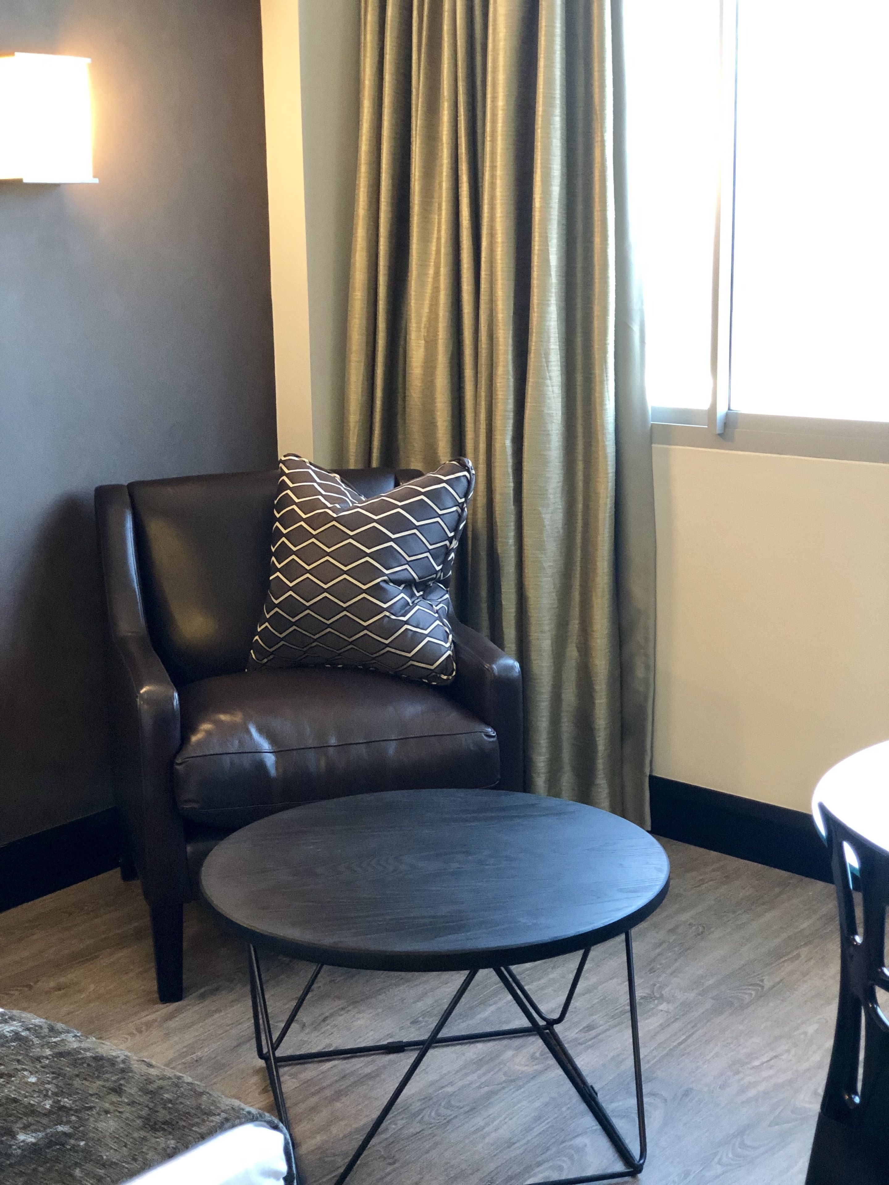 A dark leather armchair with a patterned pillow, a round table with a metal frame, and a window with curtains.