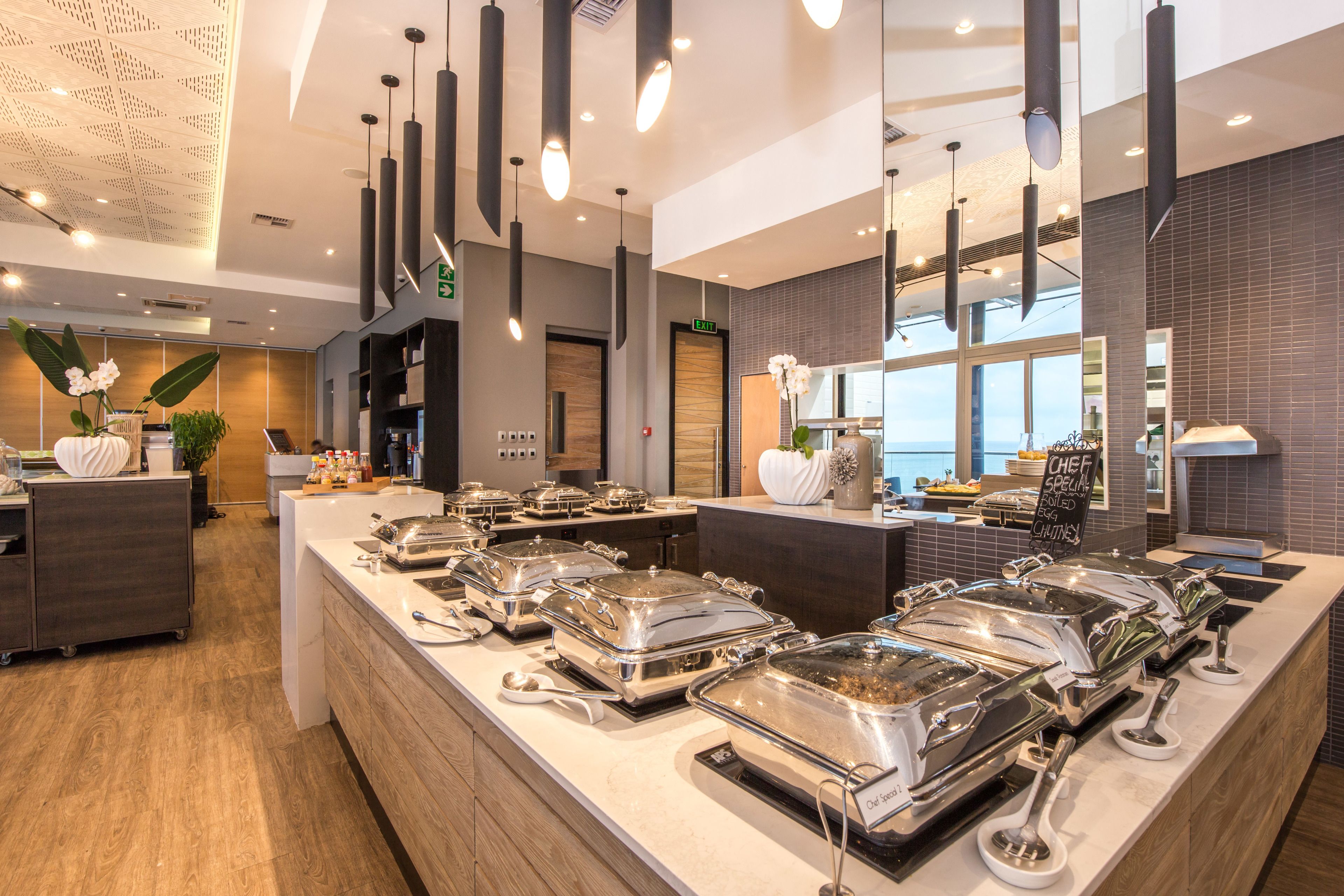 A modern hotel lobby with a buffet area featuring multiple food stations, a reception desk, and a large window offering a view of the sea.