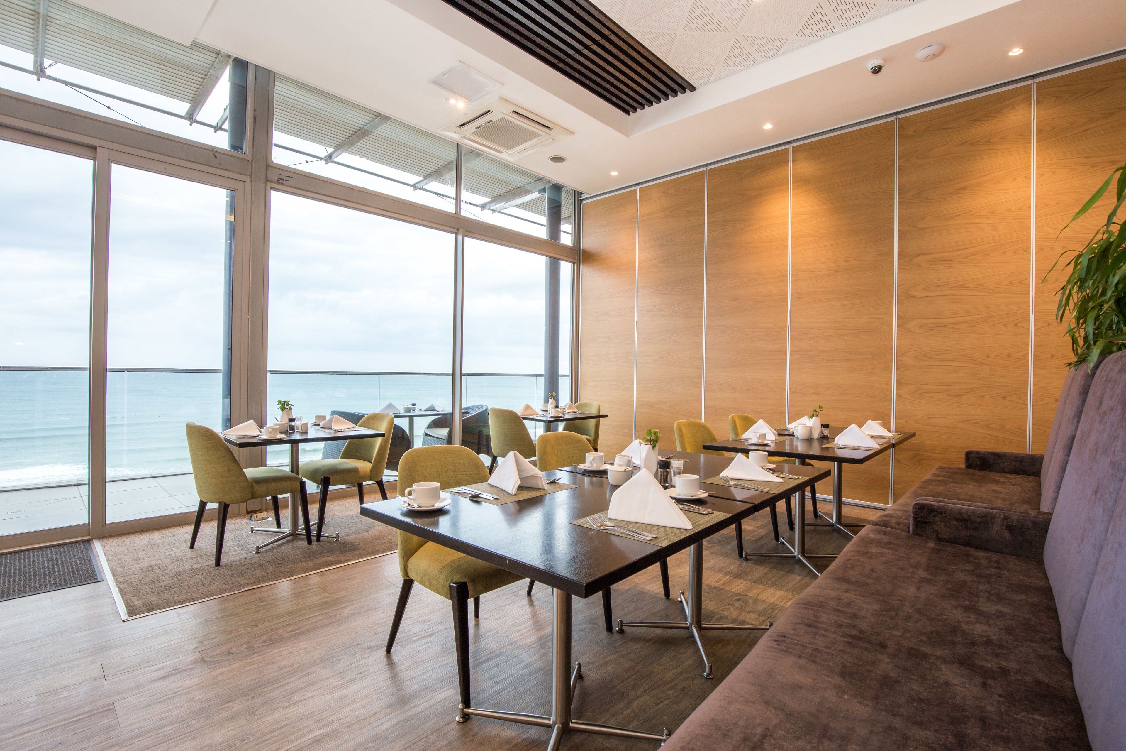 A modern conference room with large windows overlooking the sea, equipped with tables, chairs, and a sofa.
