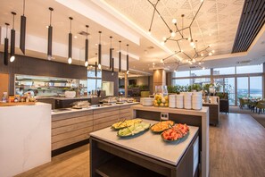 A modern hotel kitchen with a buffet setup, wooden cabinetry, and a large window offering a city view. - Belaire Suites (Durban)