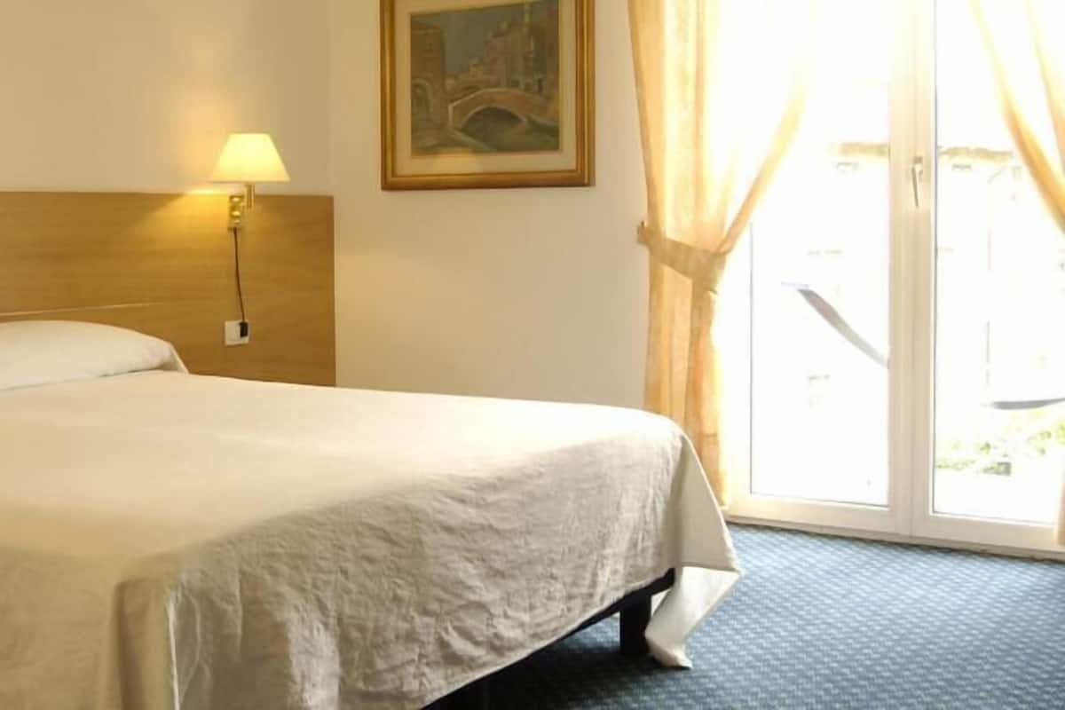 A hotel room with a bed, a nightstand with a lamp, a painting on the wall, and a view of the outdoors through a window.