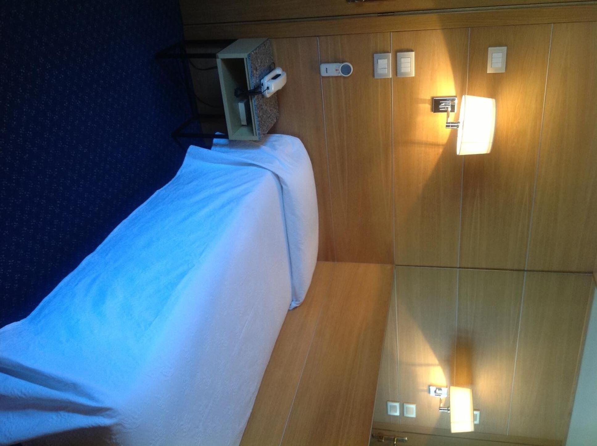 A hotel room with a bed, a wooden panel wall, and a control panel with buttons and a small screen.