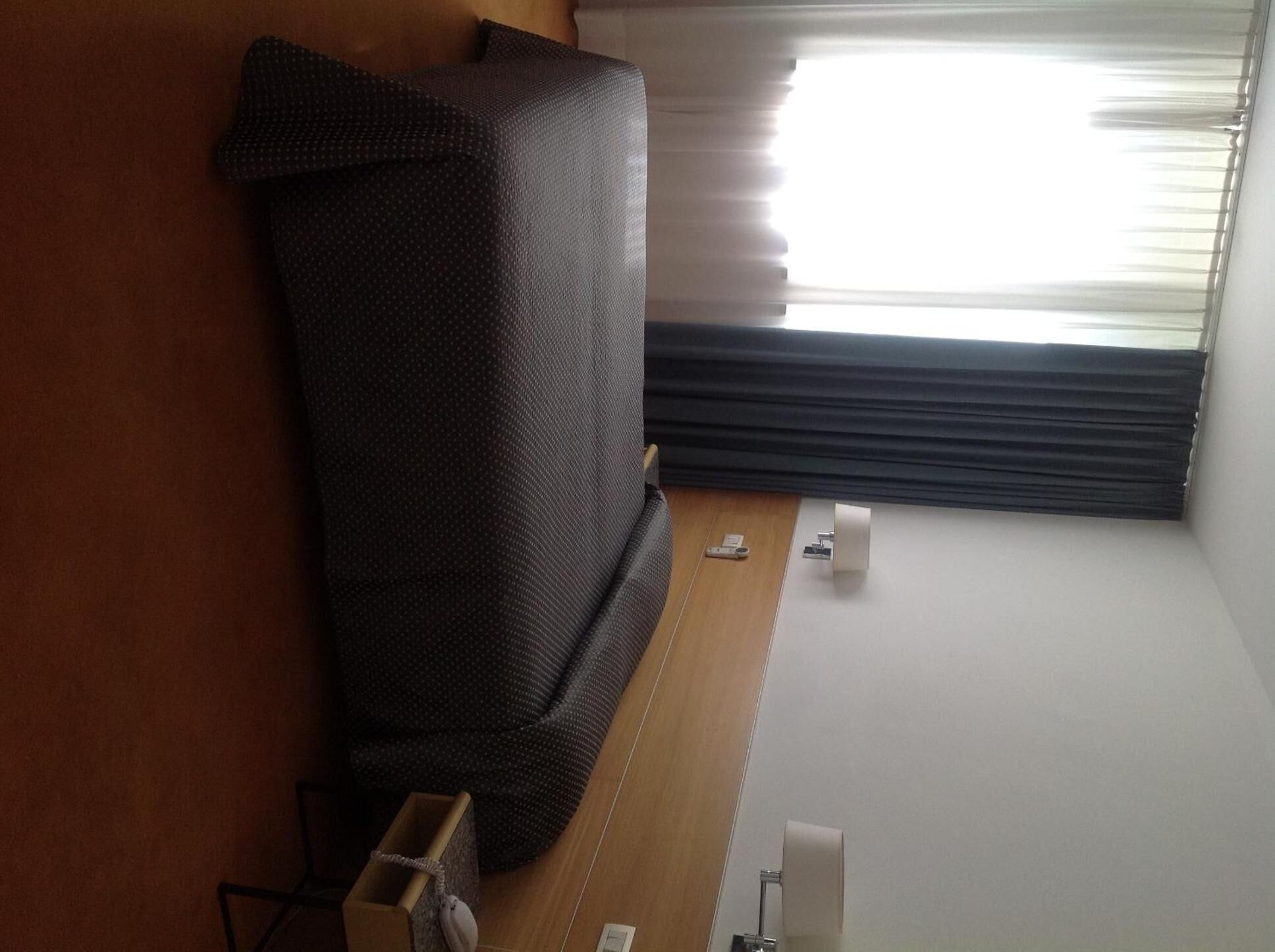 A room with a bed, a window with blinds, a small table, and a wall-mounted shelf.