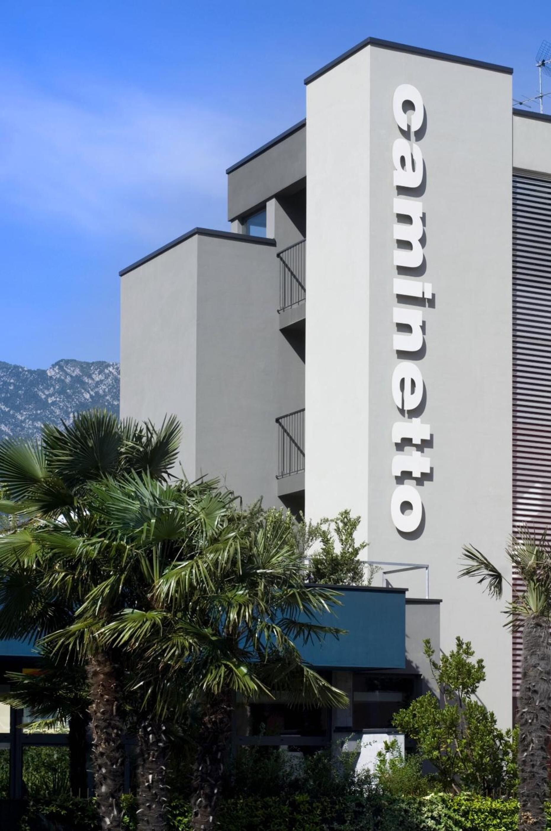A modern building with a prominent 'caminetto' sign, surrounded by lush greenery and mountains in the background.