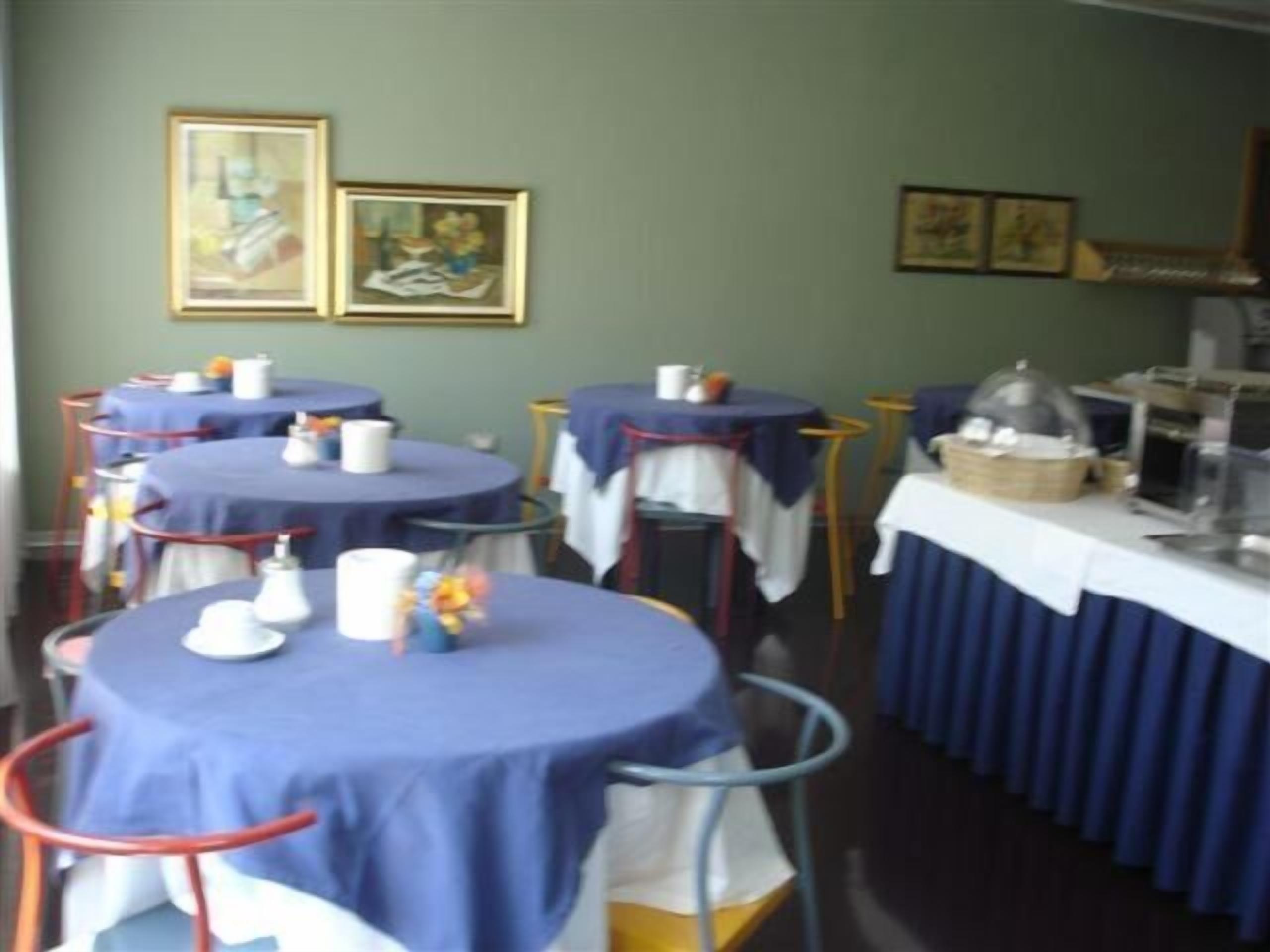 Un espace repas avec des tables rondes recouvertes de nappes bleues, des assiettes blanches et des tasses. Des tableaux encadrés ornent le mur, et un buffet est installé sous une cloche de verre.