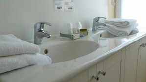 A bathroom with a white sink, two faucets, a mirror, and a towel rack with folded towels. - LOGIS HOTEL RESTAURANT DU COMMERCE (Pont-d'Ouilly)