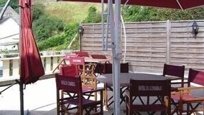 An outdoor seating area with tables and chairs, a red umbrella, and a wooden fence in the background. - LOGIS HOTEL RESTAURANT DU COMMERCE (Pont-d'Ouilly)
