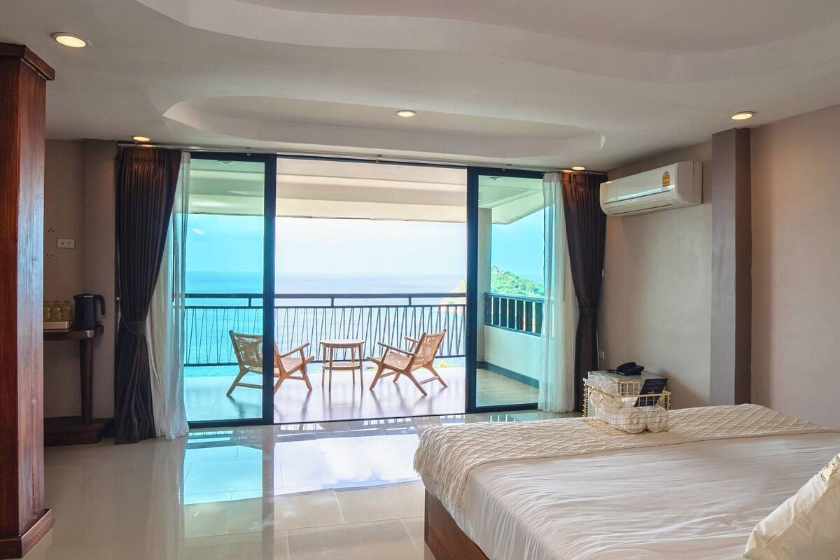 A hotel room with a large bed, a balcony with chairs, and a view of the ocean.