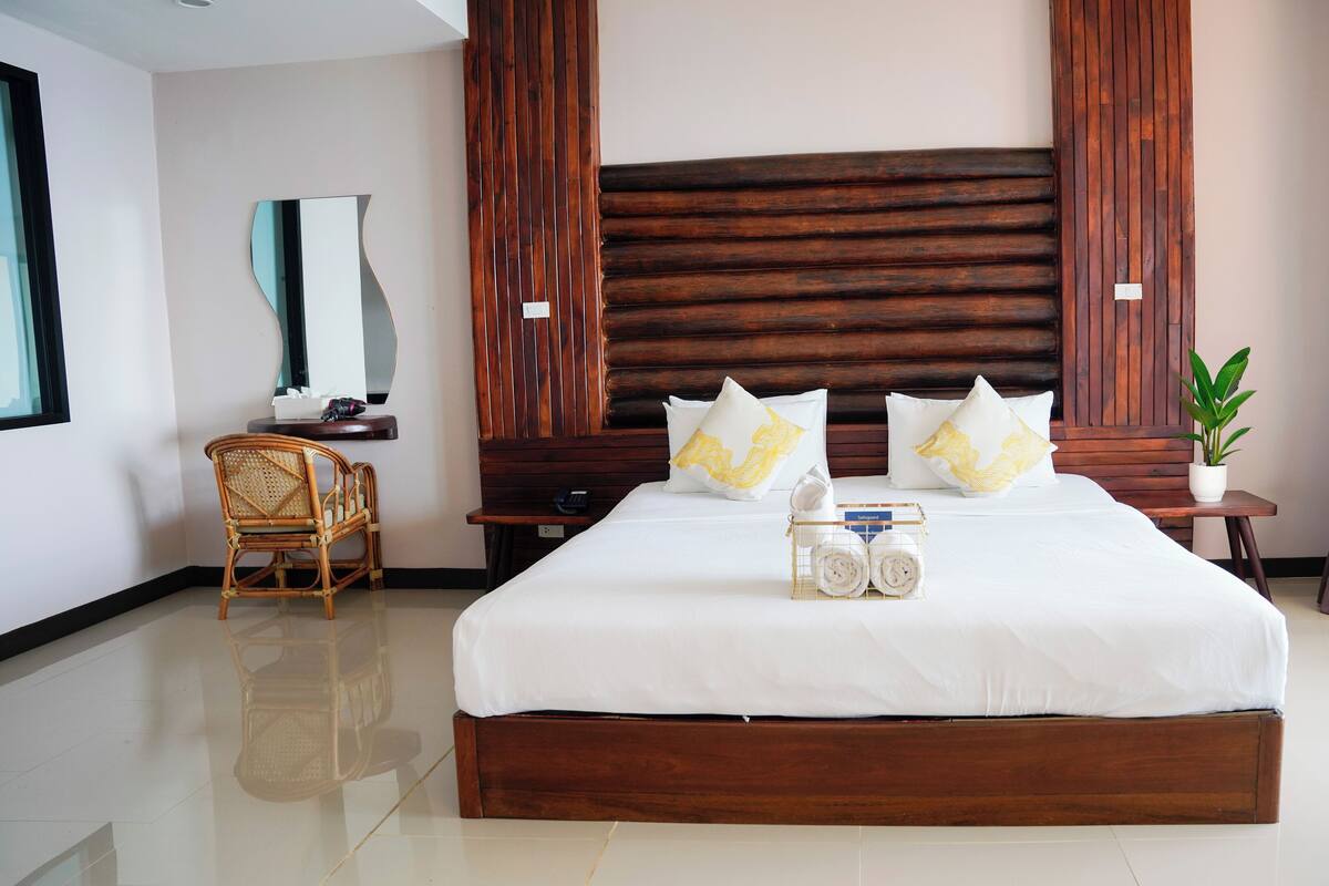A large bed with a wooden headboard, white bedding, and decorative pillows. There is a small bedside table with a plant, a chair, and a mirror on the wall.