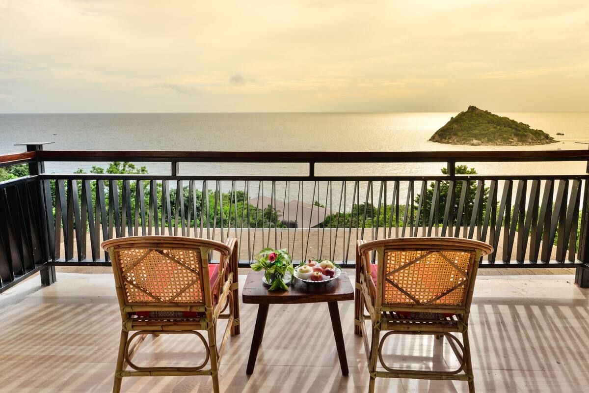 A balcony with wicker chairs and a table, overlooking a calm sea and a small island.