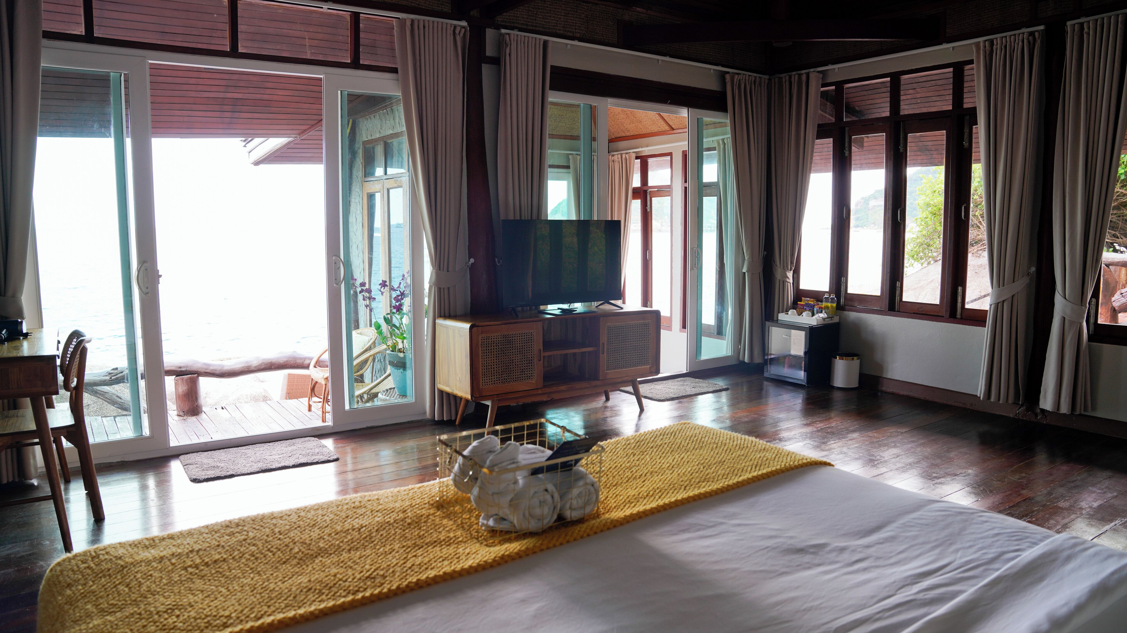 A hotel room with a bed, a television, a small table with a basket of towels, and a view of the outdoors through large windows.