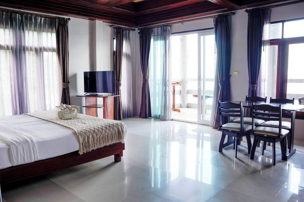 A hotel room with a large bed, a TV, a dining table with chairs, and a balcony with a view.