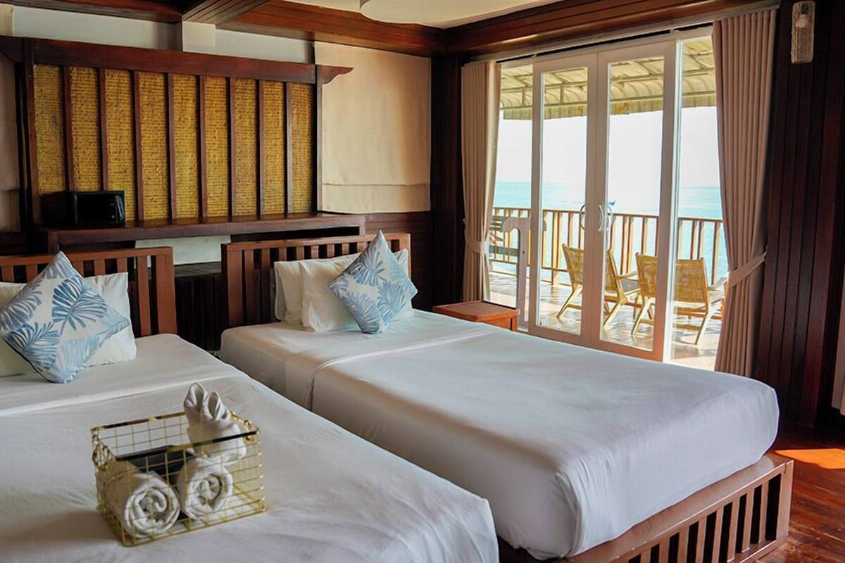 A room with two beds, a small table with towels, and a view of the ocean through large sliding doors.
