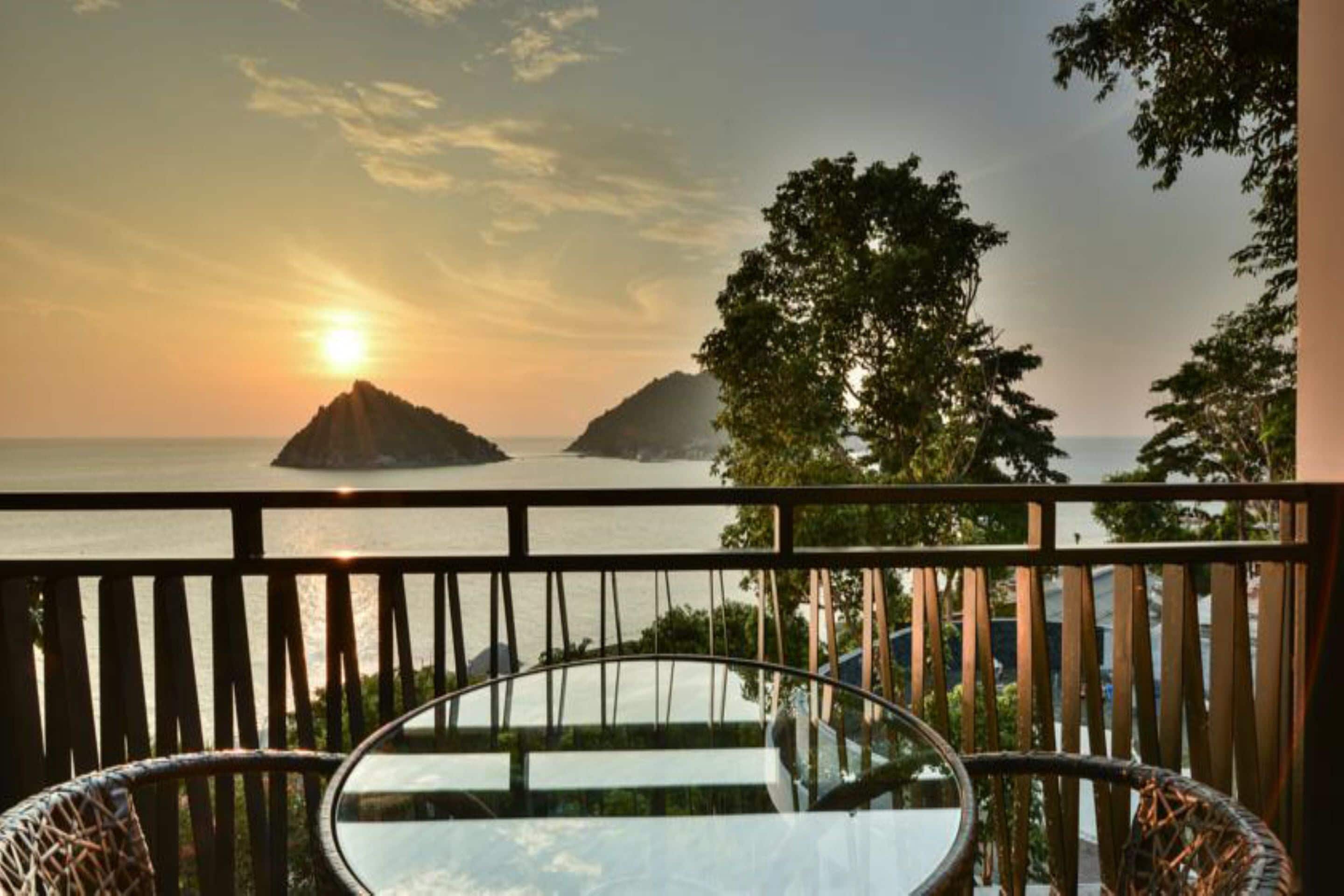 Un balcon avec une table en verre et des chaises en osier, offrant une vue sur un coucher de soleil sur la mer, avec deux îles qui se dessinent à l'horizon.