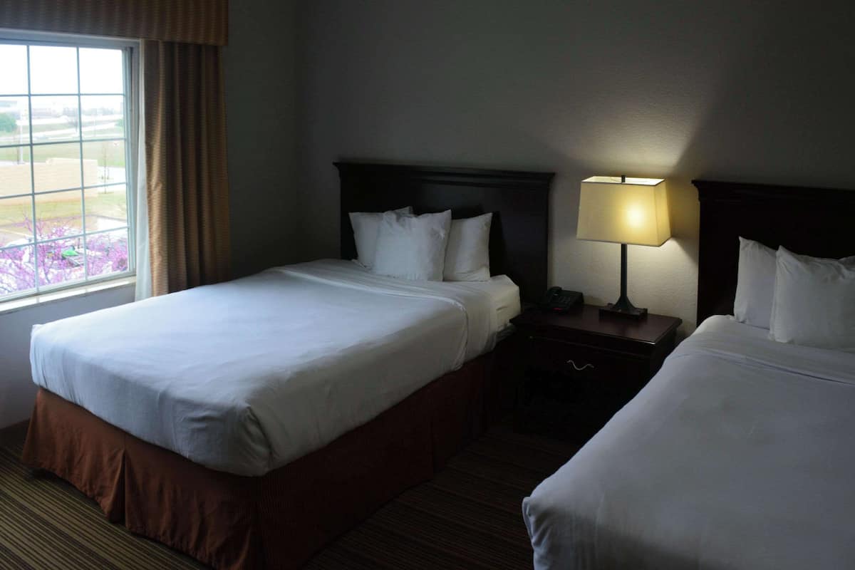 A hotel room with two beds, a nightstand with a lamp, and a window with curtains.