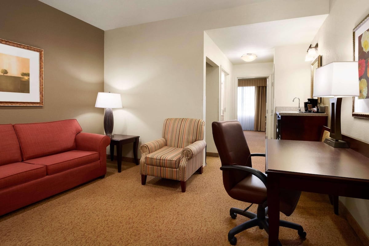 A hotel room with a red sofa, striped armchair, office chair, desk, lamp, and framed picture on the wall.