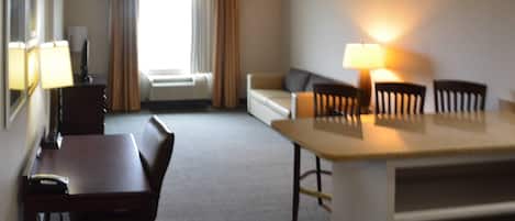 Habitación de hotel con escritorio, sillas, sofá, lámpara y ventana con cortinas.