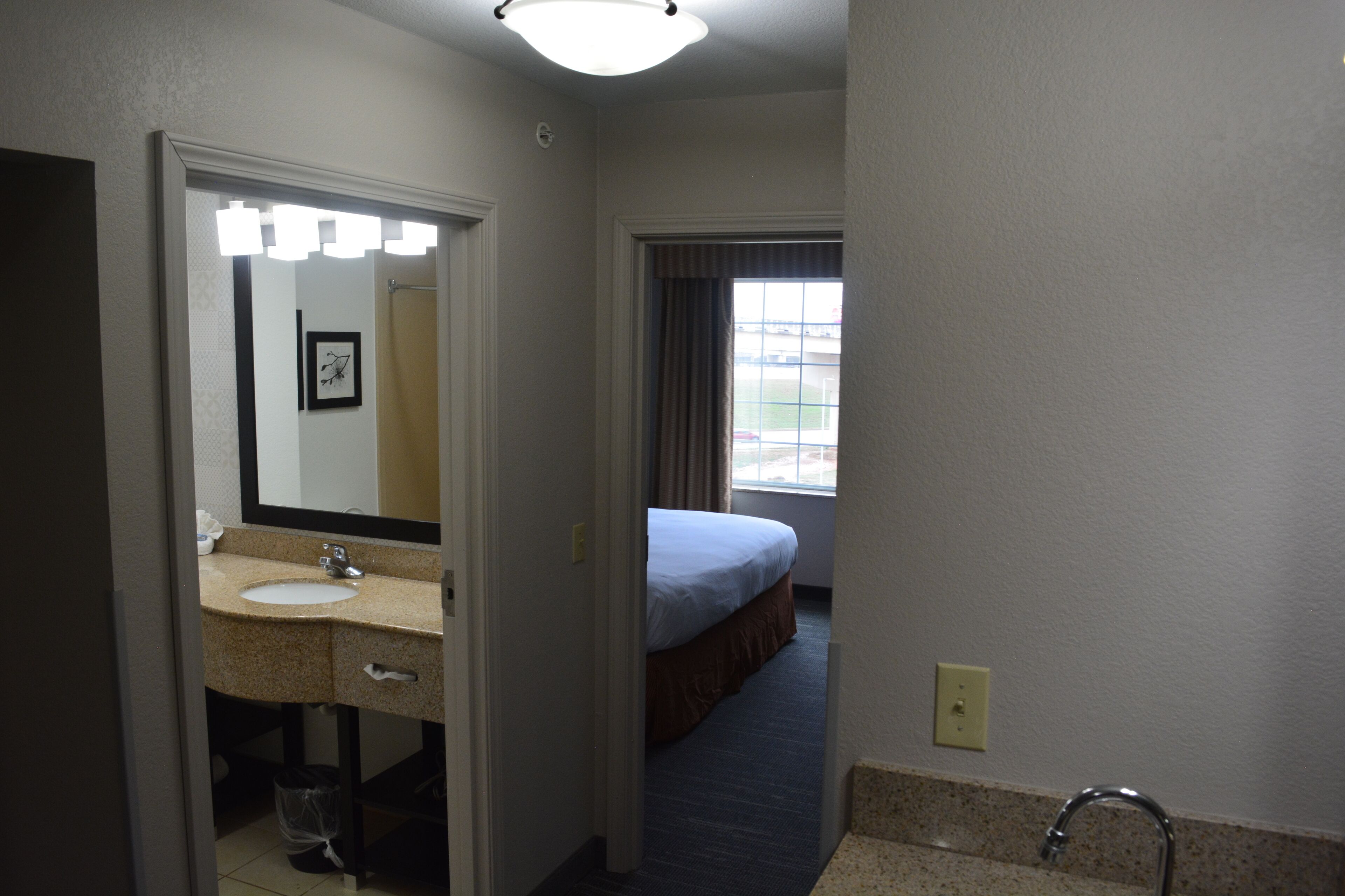 A hotel bathroom with a sink, mirror, and a bed visible through an open door.