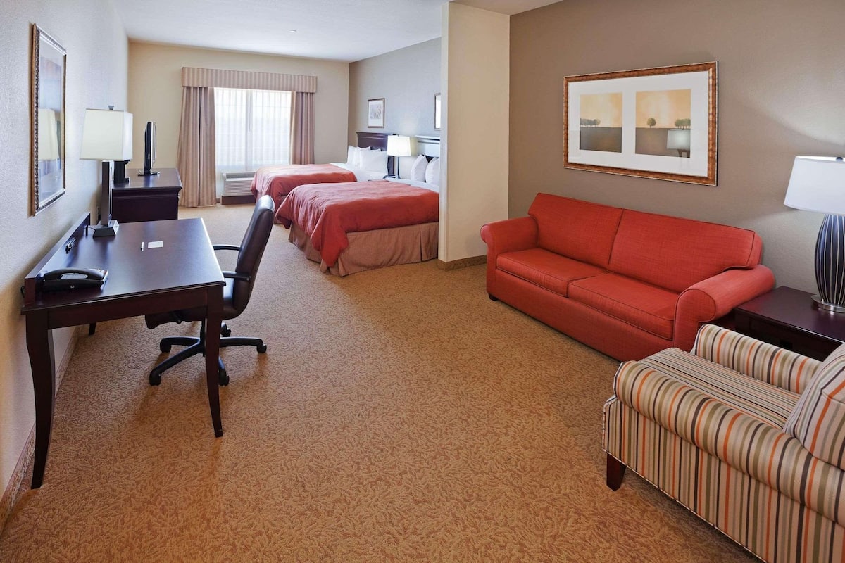 A hotel room with two beds, a desk, a red sofa, a striped armchair, and a large window with curtains.