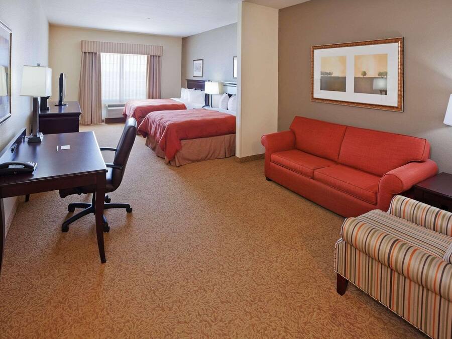 A hotel room with two beds, a desk, a red sofa, a striped armchair, and a large window with curtains.