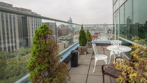 En tagterrasse med glasgelænder, hvide fletstole, et lille rundt bord og plantekasser.