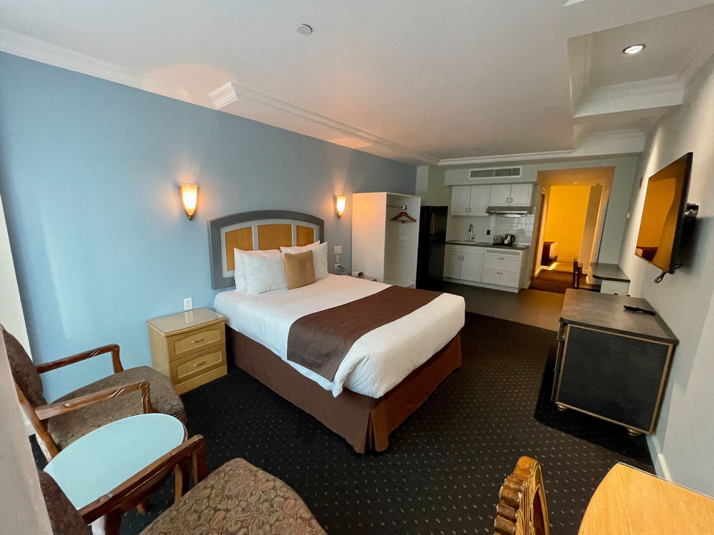 Ein Hotelzimmer mit einem großen Bett, Nachttischen, einem Schreibtisch, einem Sessel und einer Küchenzeile sowie einem Fernseher.