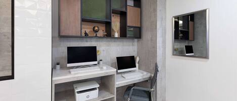 Un espacio de oficina moderno con dos escritorios, cada uno con una computadora y teclado, y una silla. Estanterías inferiores albergan varios objetos, incluyendo una impresora y adornos.