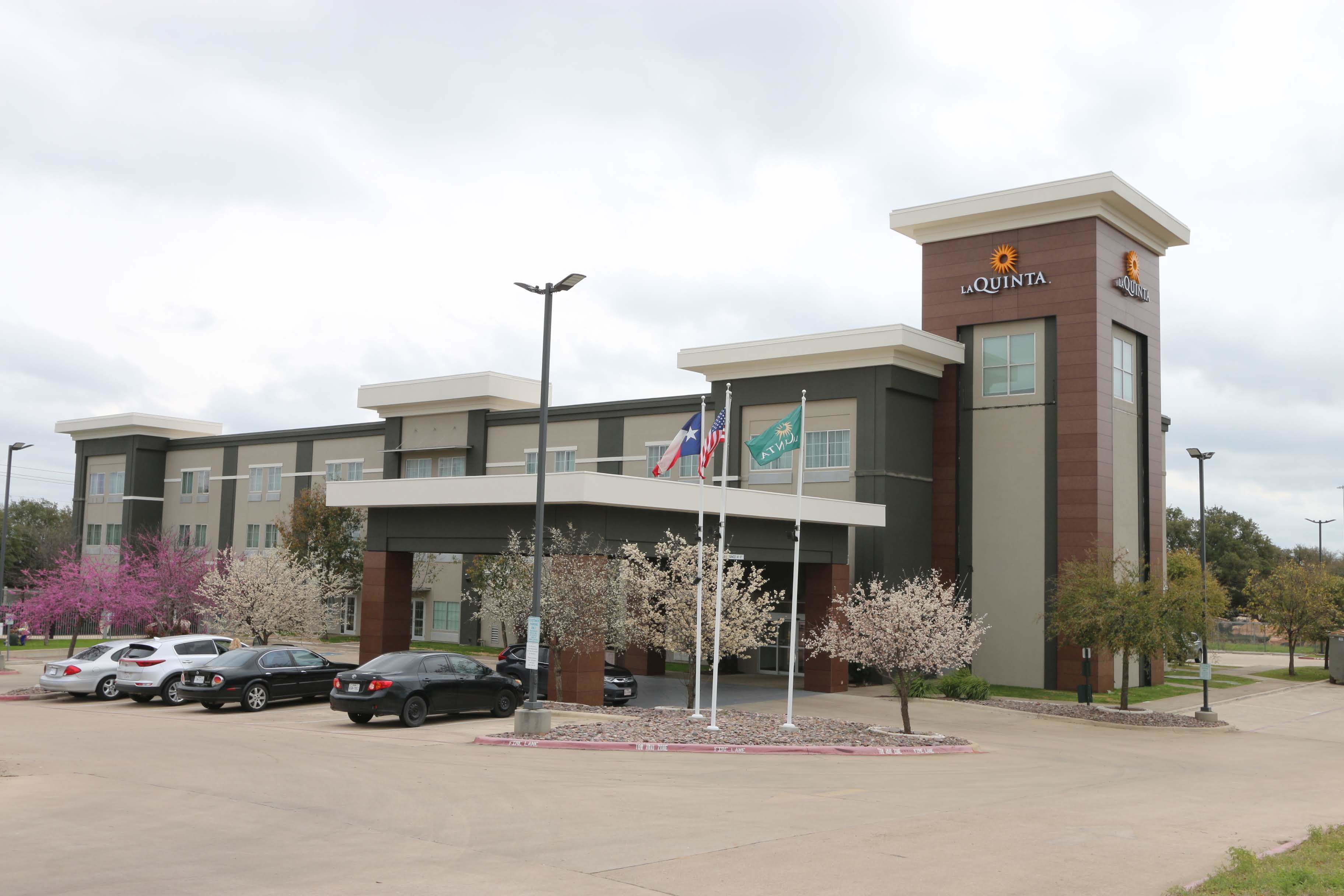 A hotel with a modern design, featuring a prominent 'La Quinta' sign, multiple flags, and a parking lot with several cars.