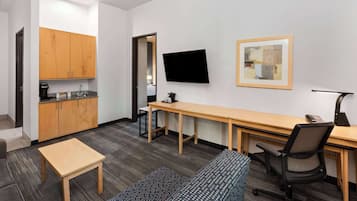 Quarto de hotel compacto com cozinha compacta, uma cama de solteiro, escrivaninha com cadeira, televisão e quadro na parede.