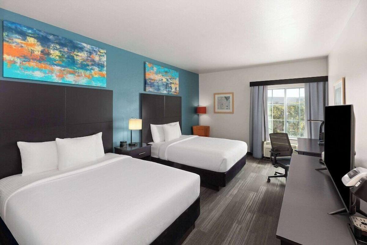 Habitación de hotel con dos camas, un escritorio, una silla, un televisor y obras de arte en las paredes.